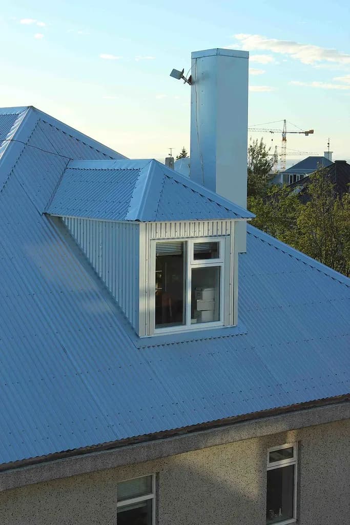 Ljóst málmþak með gluggakvisti og hvítum skorsteini.